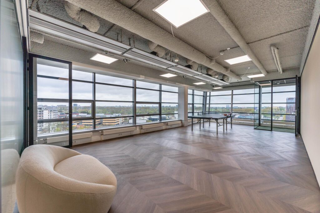 Modern office lounge with panoramic windows, a ping pong table, and a curved beige armchair on Joop Geesinkweg.